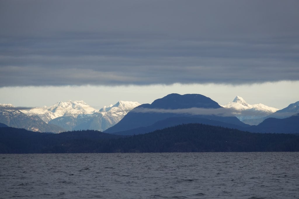 Mountain range in the distance with ocean in the foreground. The clouds in the sky are making a solid line above the moutain tops.