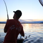 Person holding a salmon rod reeling in a fish at sunset